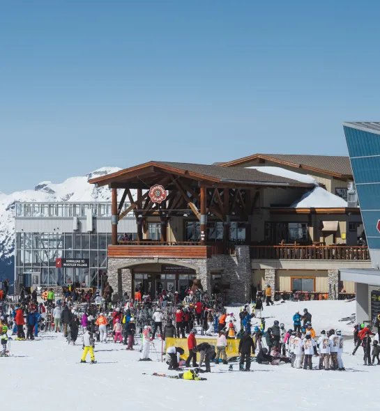 Ski lodge building with a wide, ground‑level entrance and open double doors. People in ski gear gather outside on a cleared, level entry area with railing and spacious pathways leading into the lodge.
