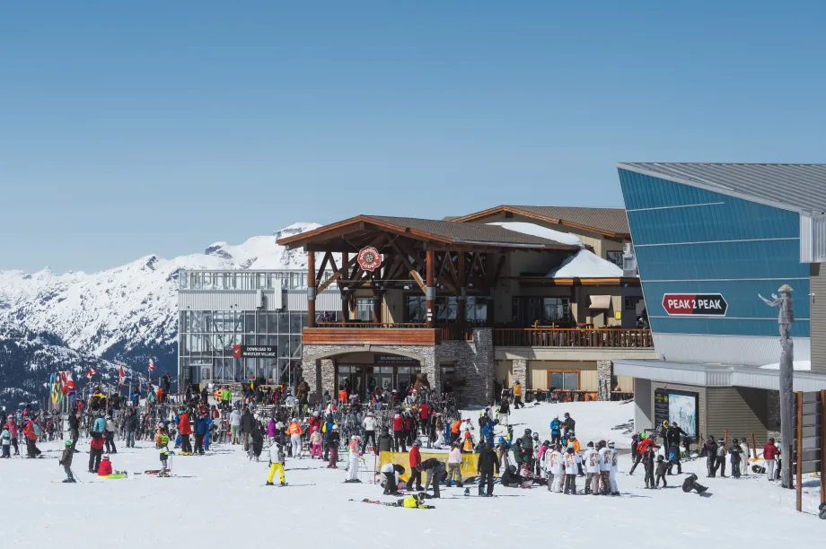 B&acirc;timent de chalet de ski avec une large entr&eacute;e de plain-pied et des portes doubles ouvertes. Des skieurs en tenue de ski se rassemblent &agrave; l'ext&eacute;rieur, sur une aire d'entr&eacute;e d&eacute;gag&eacute;e et plane avec une rambarde et de larges all&eacute;es menant au chalet.